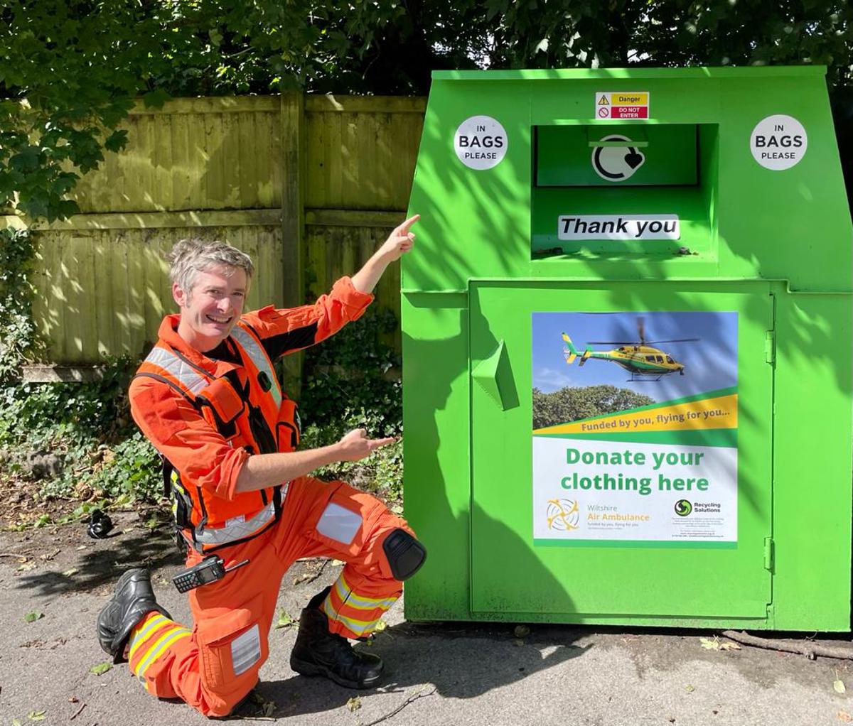 A doctor wearing an orange flight suit, crouched and pointing at a WAA green clothing bank