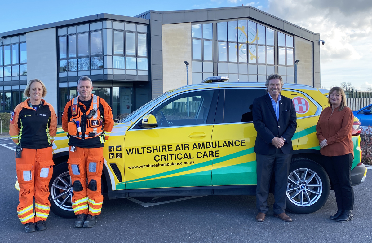 Two aircrew wearing orange flight suits posing with a yellow and green response car, with a man in a suit and woman in shirt