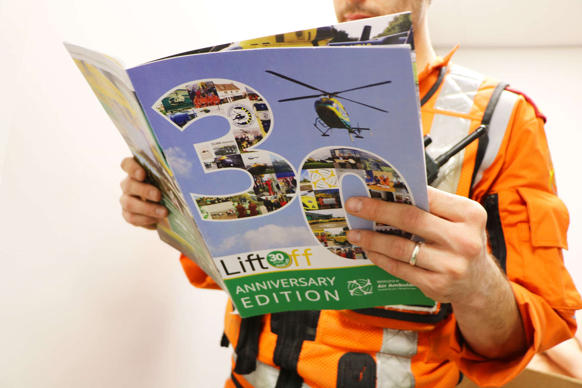 A critical care paramedic wearing an orange flight suit and reading the 30 years anniversary edition of Lift Off magazine.