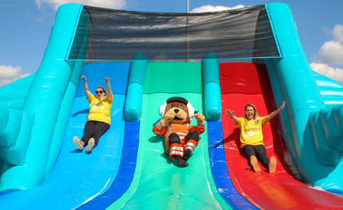 Wilber, bear mascot, sliding down an inflatable slide with two women wearing yellow Wiltshire Air Ambulance t-shirts