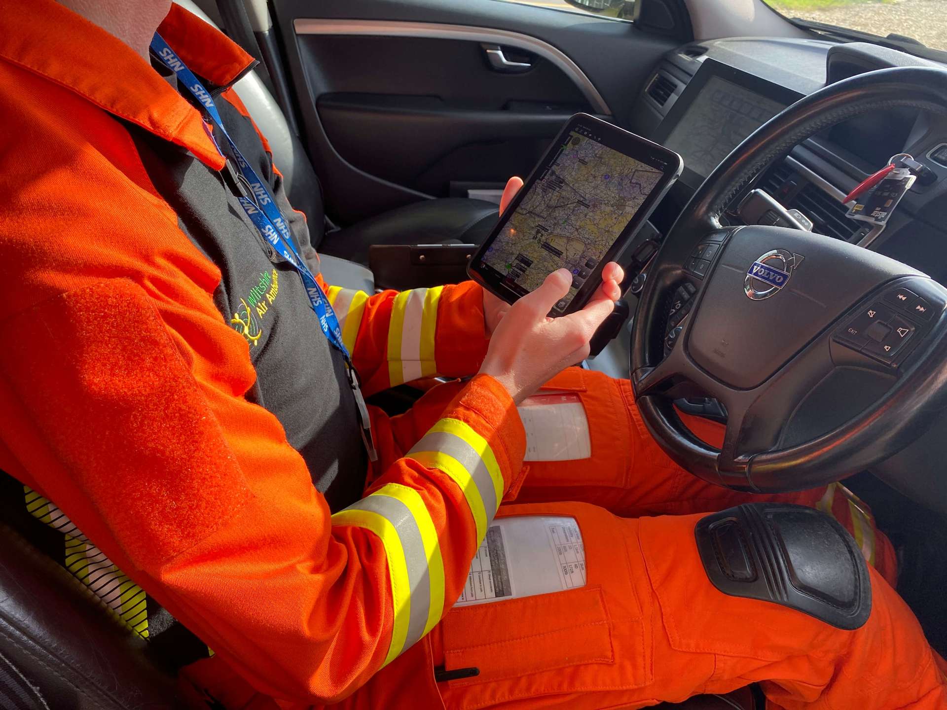 A paramedic wearing an orange flight suit sat in the drivers seat of the critical care car. They are holding an iPad which shows an image of a map.