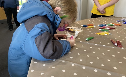 A boy wearing a blue and black jacket, with green glitter face paint, decorating a biscuit