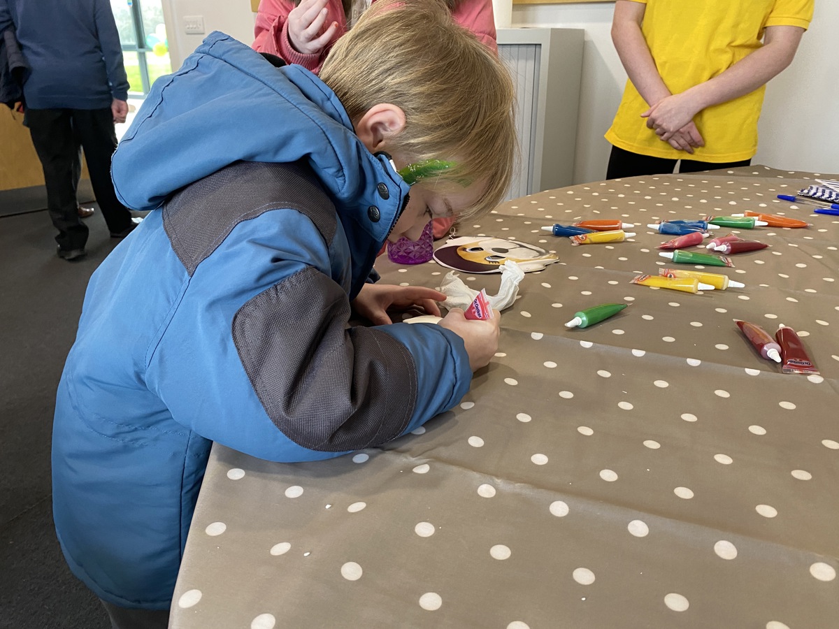 A boy wearing a blue and black jacket, with green glitter face paint, decorating a biscuit